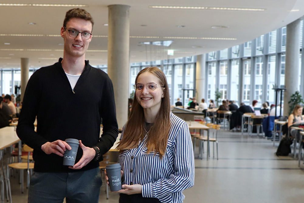 Matilda (Studentin der Biotechnologie an der Ernst-Abbe-Hochschule Jena) und Tobias (Student der Pharmazie an der Friedrich-Schiller-Universität Jena) mit ihren Gewinnsprüchen auf den Mehrwegbechern bei der Übergabe in der Mensa Ernst-Abbe-Platz in Jena